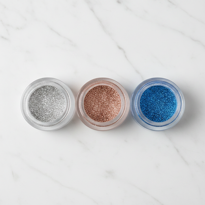 Three clear glass glitter eyeshadow jars in silver, rose gold and electric blue arranged on marble surface