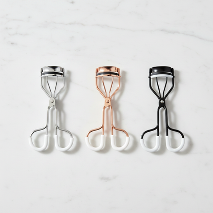 Three eyelash curlers in silver chrome, rose gold and matte black laid flat on marble surface
